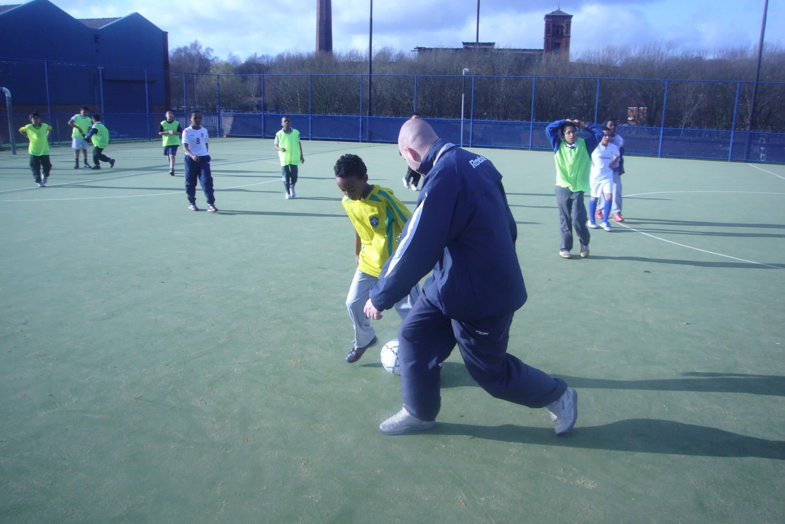 A BSCA coach playing football with young people on an outdoor pitch in Bolton